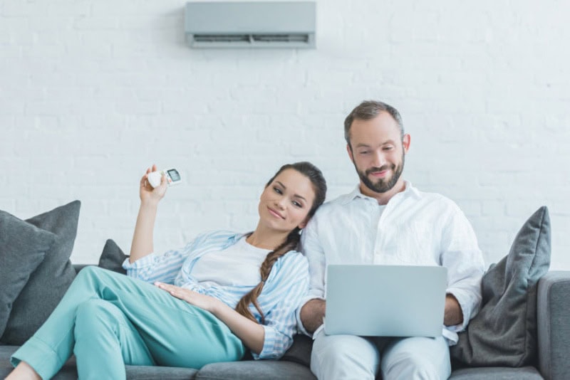 Happy couple sitting on couch looking at laptop while female uses remote to turn on ductless mini split.