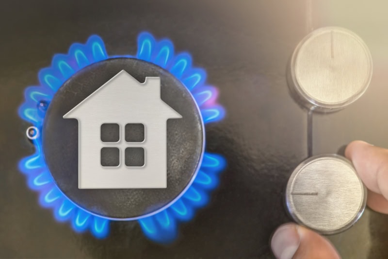 A lit gas stove burner with bright blue flames ringed around a central cap featuring a white house-shaped icon with a chimney and four windows.