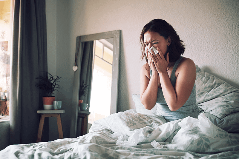 Woman in bed blowing her nose due to allergies. Representing information on your AC helping.