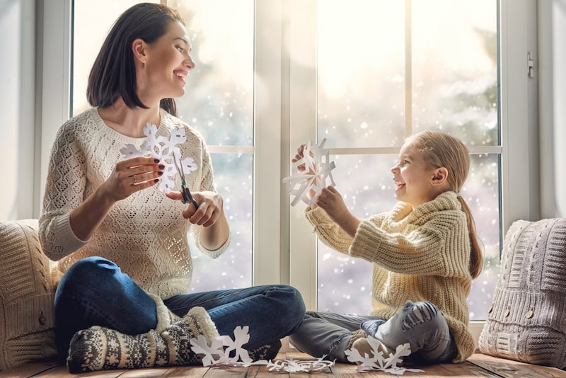 Happy loving family sitting by the window and making paper snowflakes for decoration windows. Mother and child creating decorations. A new furnace is needed in their home.